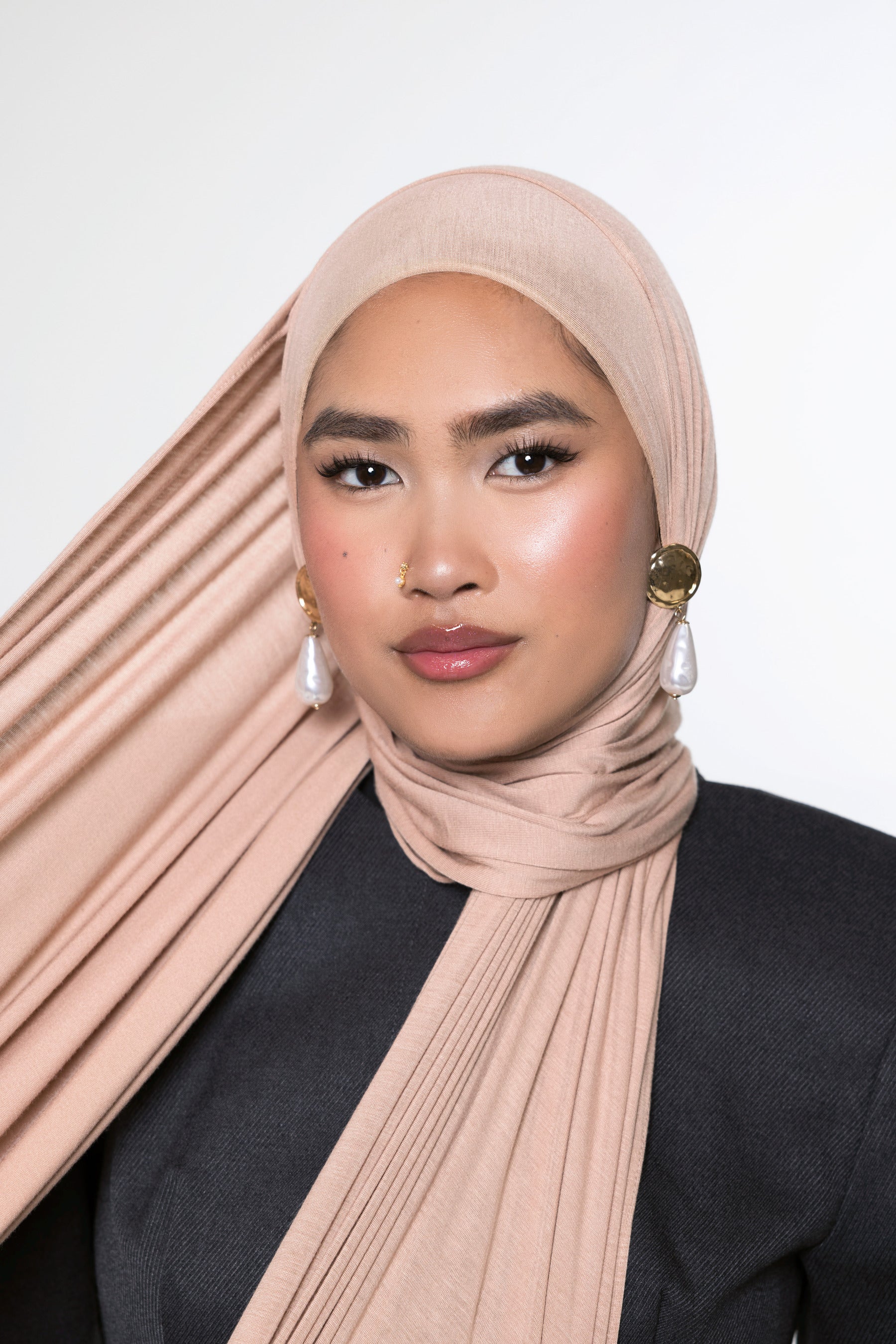 A woman wearing a pleated, draped hijab in a dusty blush pink color, paired with a black top and gold and pearl earrings, set against a plain light gray background.