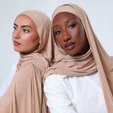 Two women wearing beige hijabs and white tops, posed back-to-back against a light gray studio background.