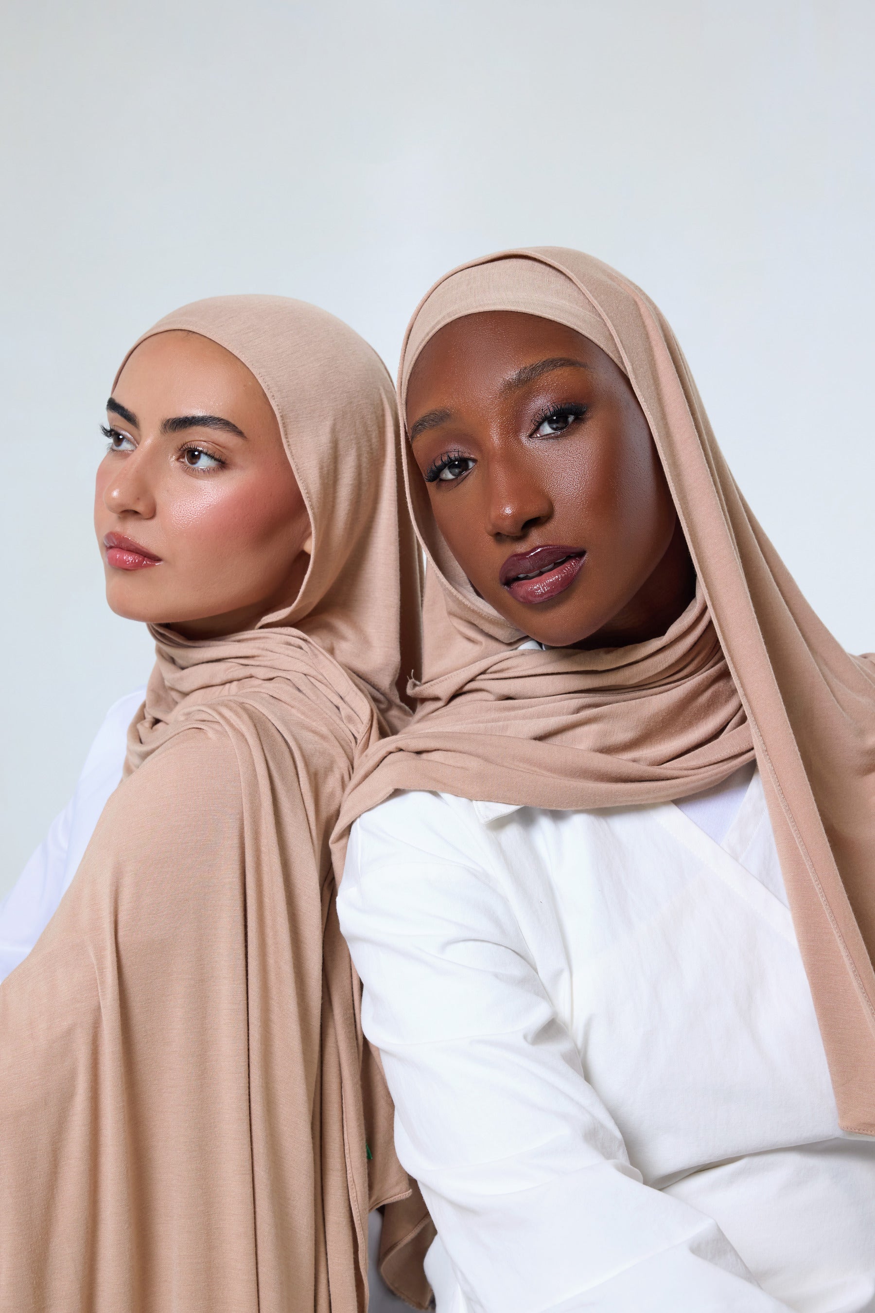 Two women wearing beige hijabs and white tops, posed back-to-back against a light gray studio background.