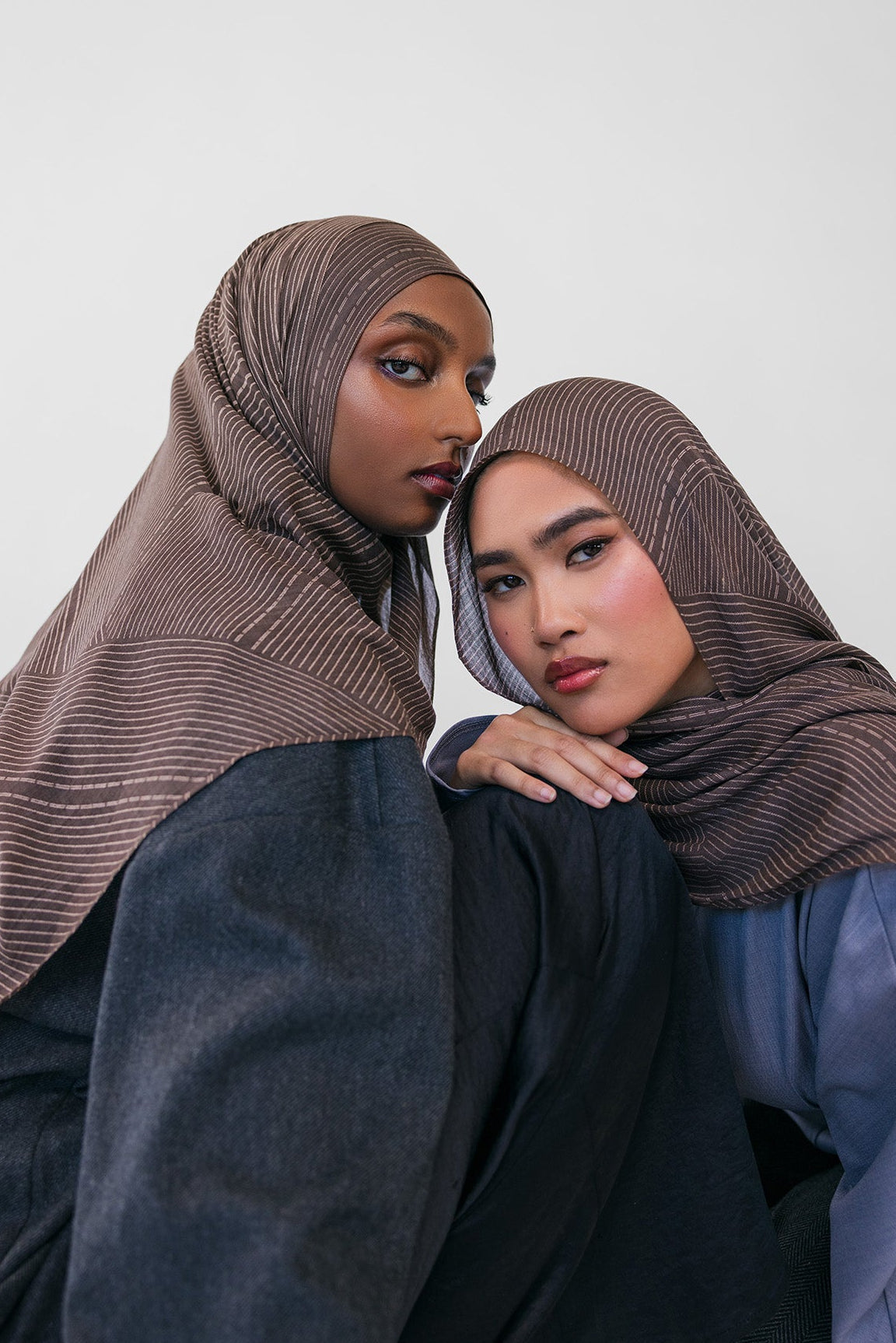 Two women wearing brown striped hijabs and dark clothing against a white background