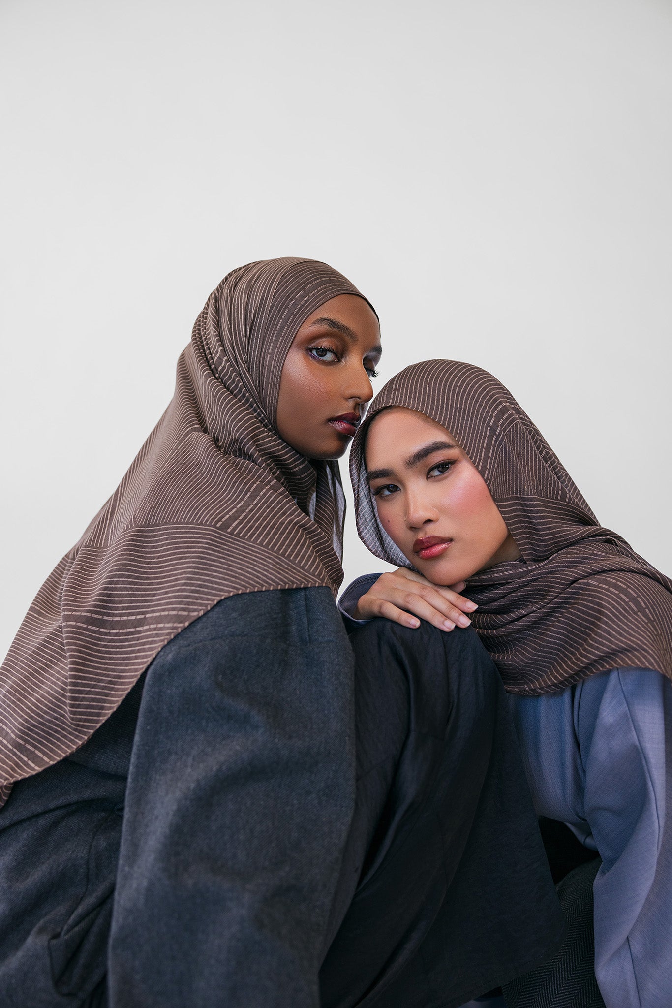 Two women wearing brown striped hijabs and dark clothing against a white background