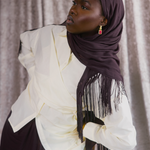 A woman wearing a dark plum purple hijab with fringe and a cream-colored wrap dress, posing against a textured gray curtain backdrop.
