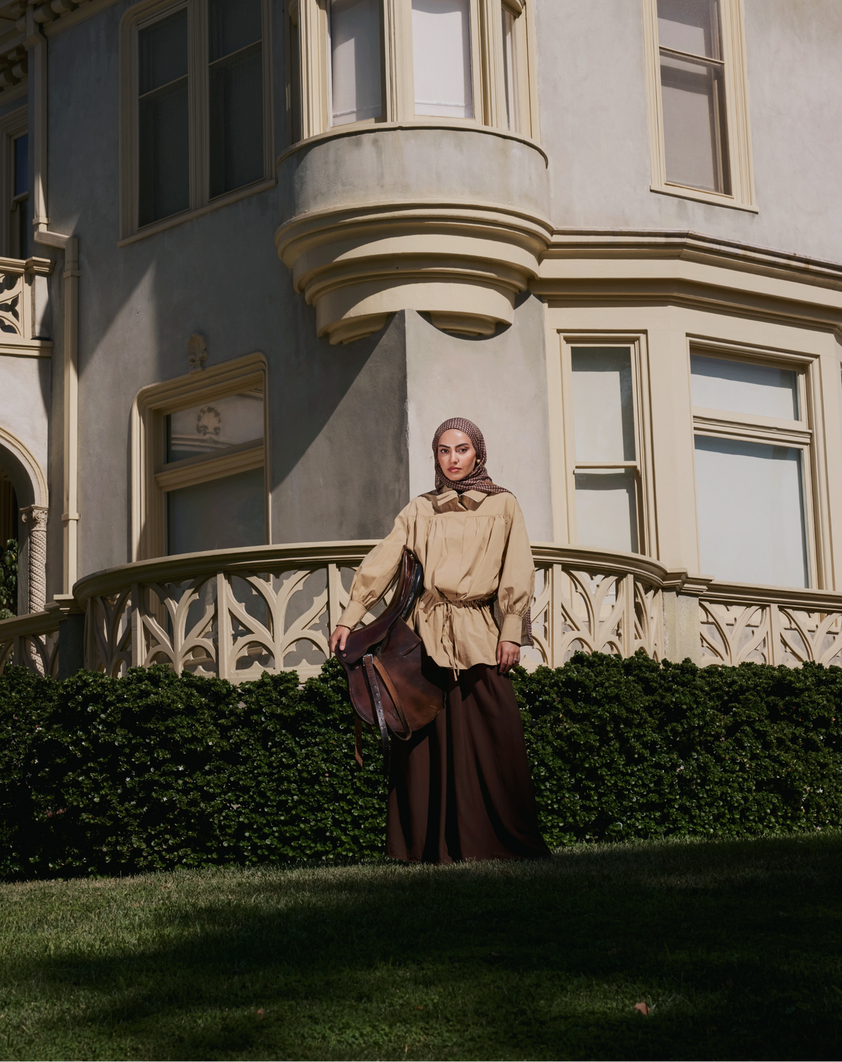 A woman wearing a vintage-style beige blouse and dark brown skirt stands on a lawn in front of a large Victorian-style mansion.