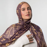 A woman wearing a brown and gold patterned hijab and a white ribbed sweater, photographed against a light gray studio background.