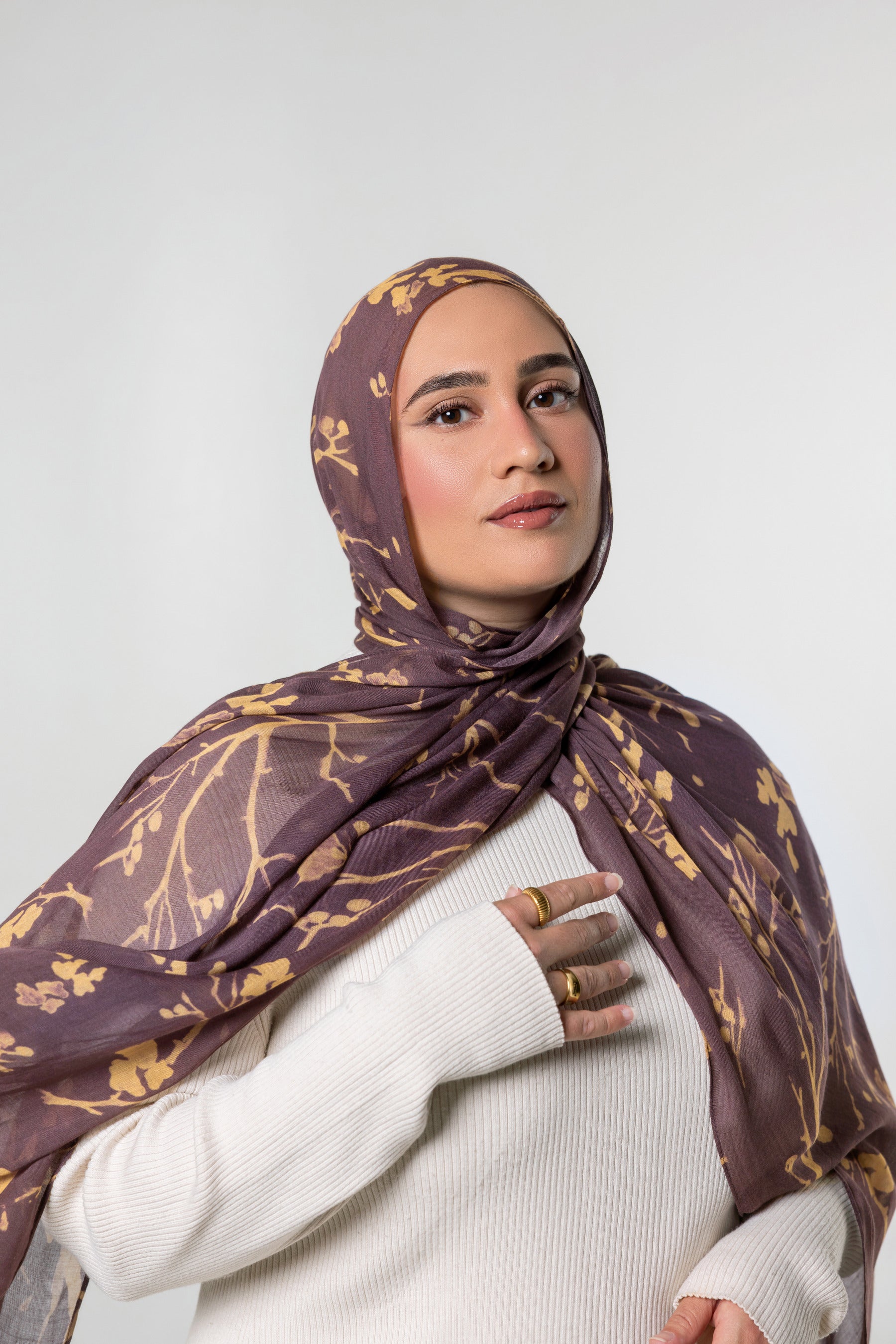 A woman wearing a brown and gold patterned hijab and a white ribbed sweater, photographed against a light gray studio background.
