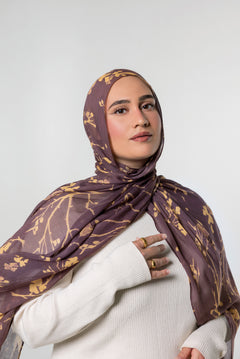A woman wearing a brown and gold patterned hijab and a white ribbed sweater, photographed against a light gray studio background.