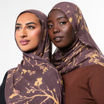 Two women wearing matching brown and gold patterned hijabs, posed against a clean light gray studio background.