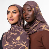 Two women wearing matching brown and gold patterned hijabs, posed against a clean light gray studio background.