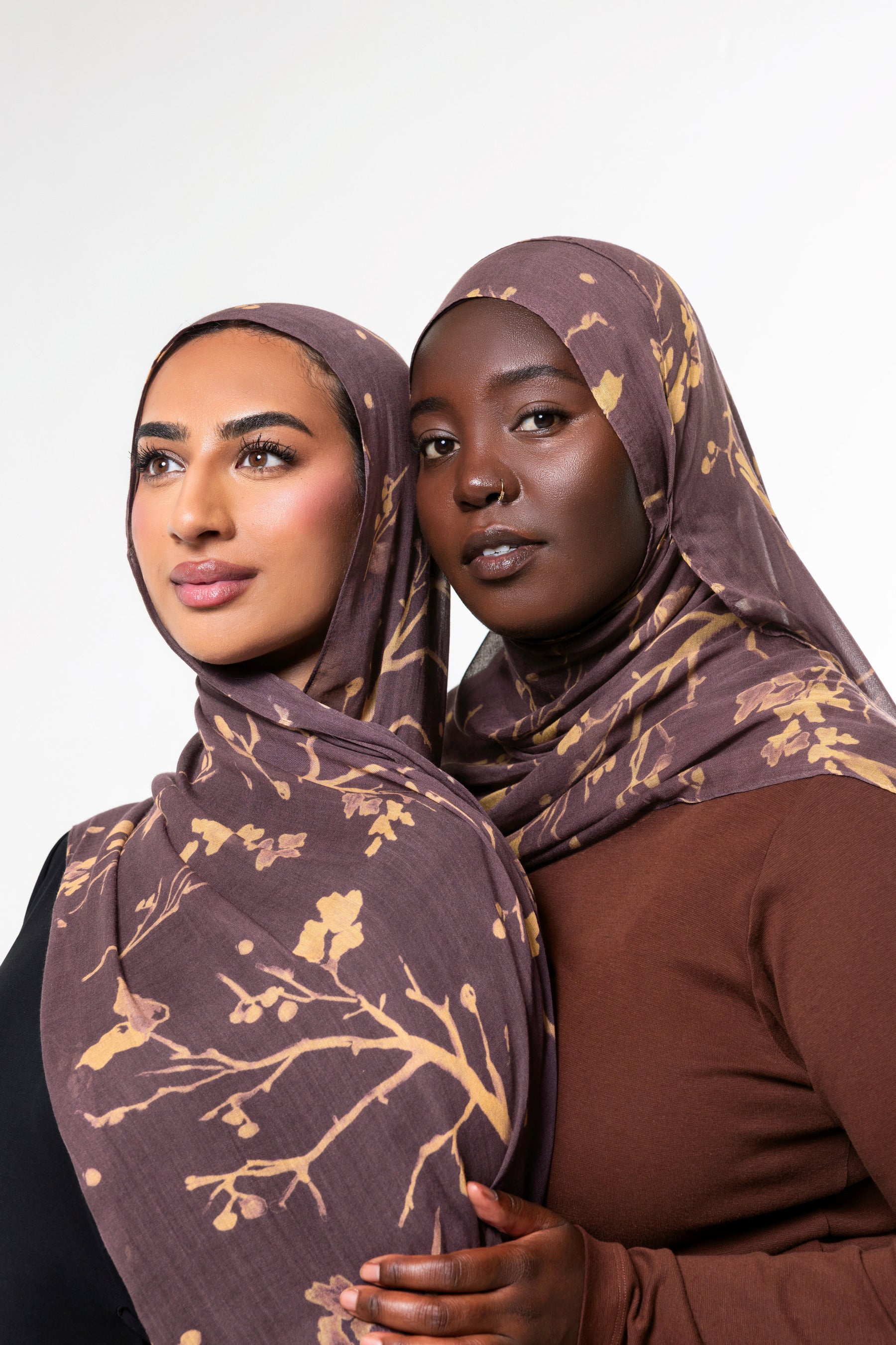 Two women wearing matching brown and gold patterned hijabs, posed against a clean light gray studio background.