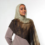A woman wearing a taupe hijab and a matching long-sleeve dress, draped with a sheer brown and beige patterned shawl over her shoulders, set against a plain light gray studio background.