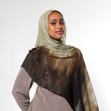 A woman wearing a taupe hijab and a matching long-sleeve dress, draped with a sheer brown and beige patterned shawl over her shoulders, set against a plain light gray studio background.