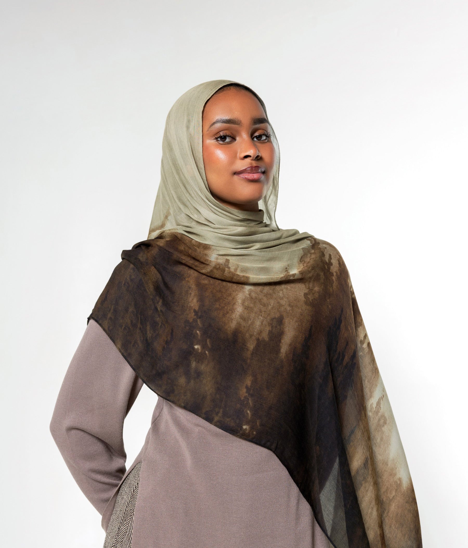 A woman wearing a taupe hijab and a matching long-sleeve dress, draped with a sheer brown and beige patterned shawl over her shoulders, set against a plain light gray studio background.