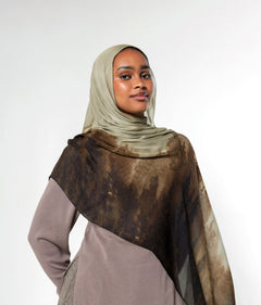 A woman wearing a taupe hijab and a matching long-sleeve dress, draped with a sheer brown and beige patterned shawl over her shoulders, set against a plain light gray studio background.