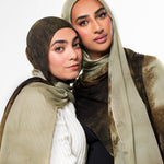 Two women wearing matching hijabs and shawls in earthy beige, brown, and white tones, posed closely against a plain light background.