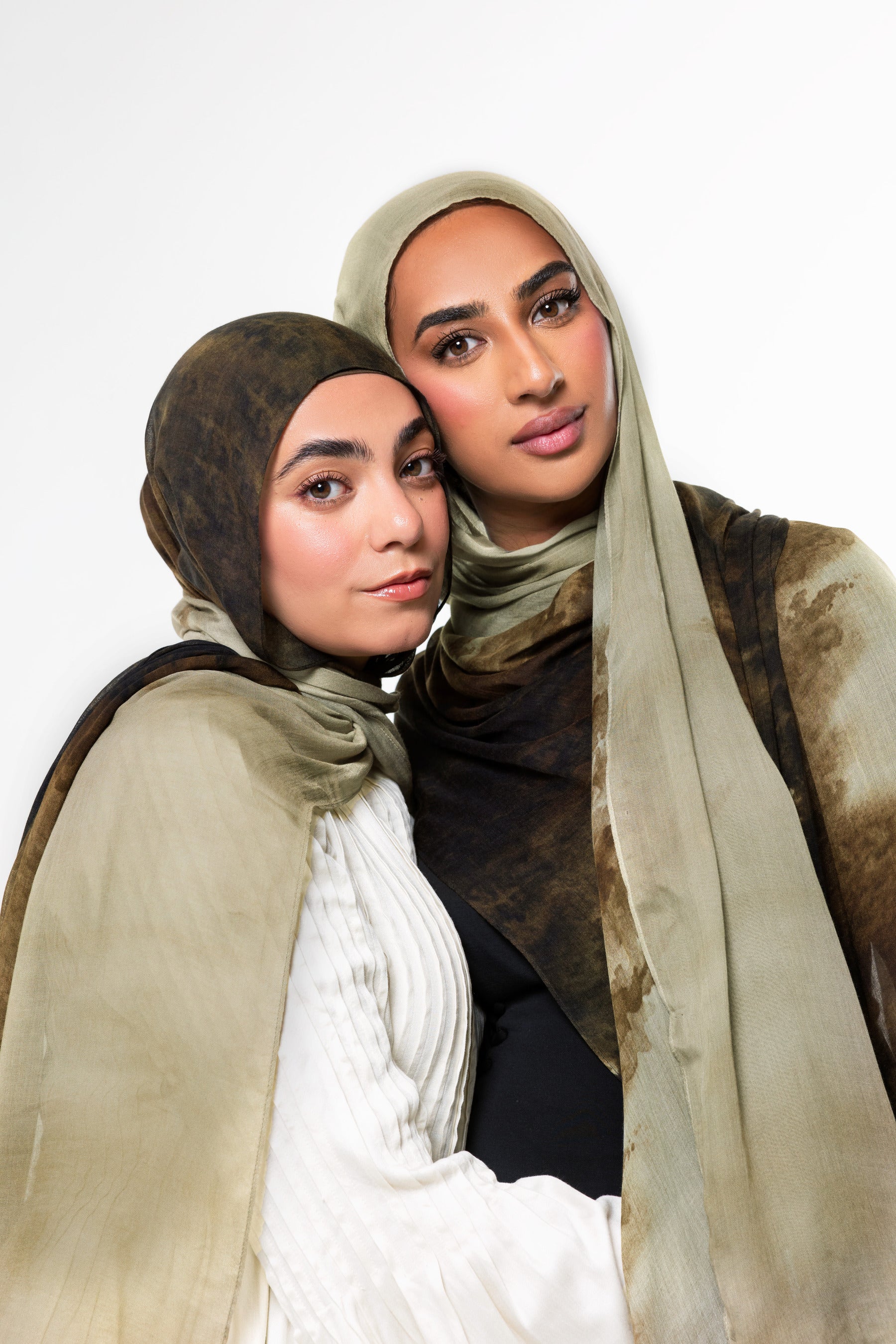 Two women wearing matching hijabs and shawls in earthy beige, brown, and white tones, posed closely against a plain light background.