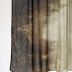 A sheer curtain panel with a mottled, abstract pattern in dark brown, charcoal gray, and beige, hanging against a plain white background.