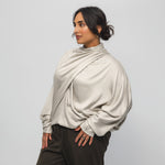 Woman wearing a beige long-sleeve top and dark pants against a light gray background