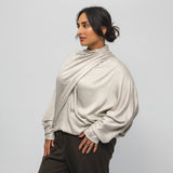 Woman wearing a beige long-sleeve top and dark pants against a light gray background