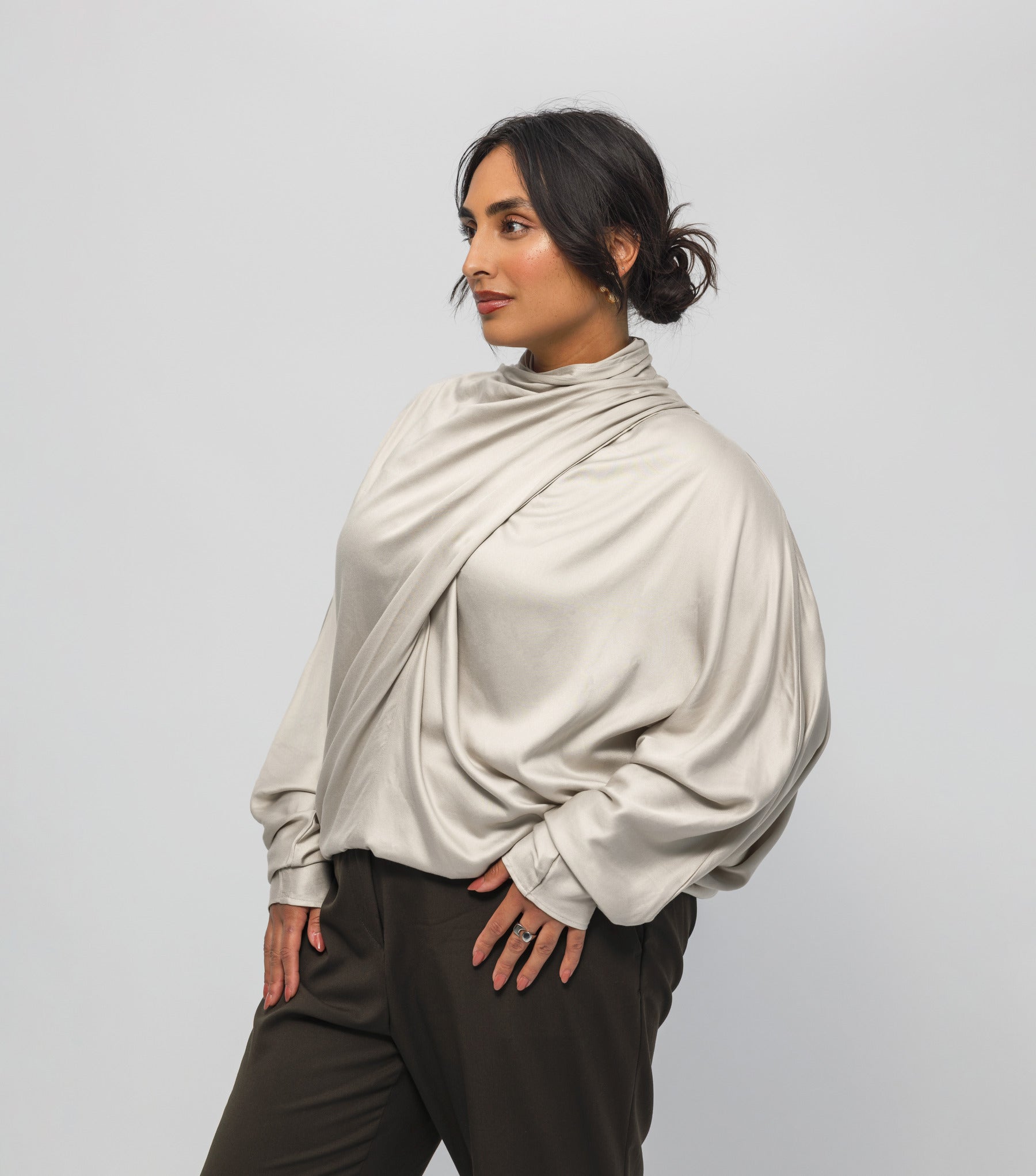 Woman wearing a beige long-sleeve top and dark pants against a light gray background