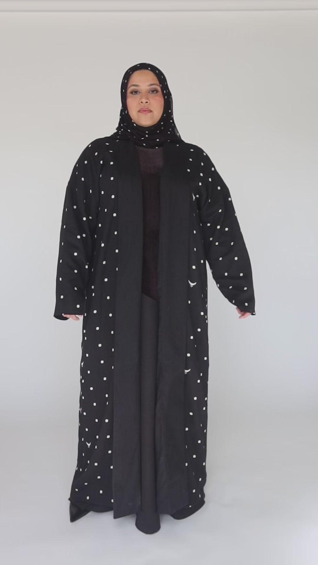 Woman wearing a long black coat with white polka dots and a matching headscarf on a gray background